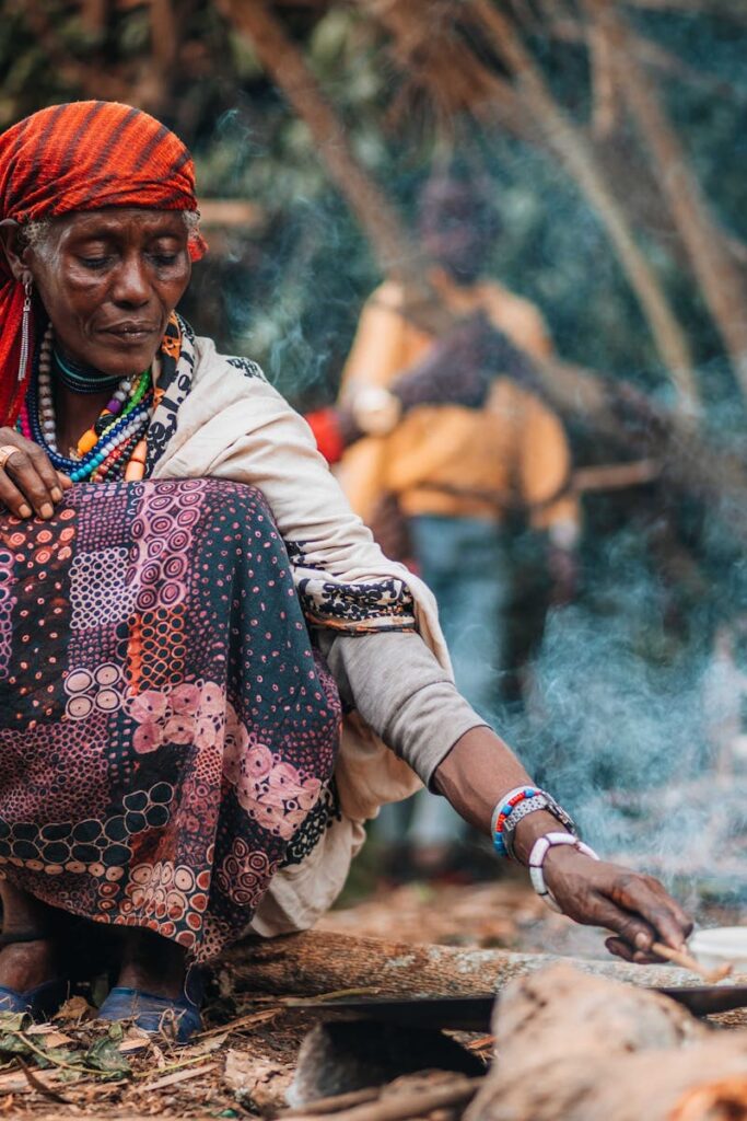 A woman cooking outdoors in a traditional African village setting with visible smoke.