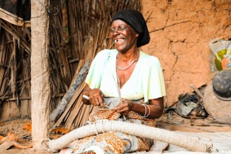 An African woman smiles as she weaves in a rural Tanzanian setting, showcasing traditional craftsmanship.