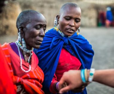 Two Maasai women adorned in traditional attire share a moment outdoors, wearing colorful beads and clothing.